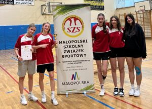 Zwycięska drużyna koszykarska dziewcząt z ZSE z pucharem i medalem za zwycięskie zawody. Drużyn pozuje z banerem Małopolskiego Szkolnego Związku Sportowego.