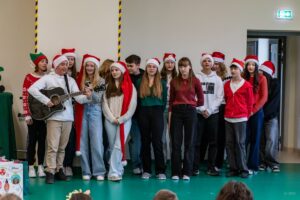 Grupa uczniów w czapkach św. Mikołaja śpiewa. Jeden uczeń gra na gitarze.