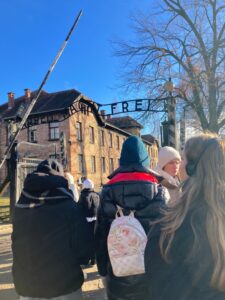 Uczniowie stoją przed wejściem do obozu Auschwitz-Birkenau w Oświęcimiu. W tle brama wejściowa.