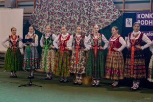 Uczennice w strojach regionalnych stojące w szeregu śpiewają piosenkę w gwarze regionalnej.