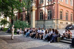 Cała grupa siedząc na schodach przy Collegium Novum słucha objaśnień Pani Przewodnik.