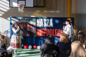 0. Na tle dekoracji z napisem Nasza Niepodległa uczennica w stroju regionalnym i uczeń deklamują wiersze.