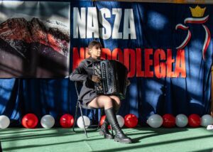 23. Na scenie na tle scenografii występuje ładna akordeonistka.