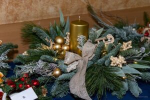 Zbliżenie na stroik , w którego środku znajduje się złota świeca a wokół niej bombki choinkowe i ozdoby też w kolorze złotym.