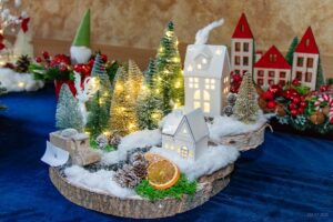 Zbliżenie na stroik złożony z białych domków, zielonych minidrzewek i ozdób świątecznych.
