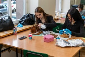 Dwie uczennicy ozdabiają pierniki, jedna skleja piernikowy domek druga przygotowuje element z lukrem.