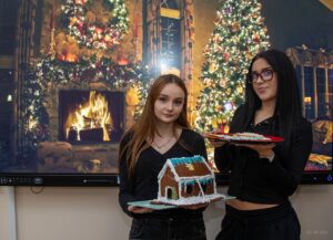 Jeszcze jedno zdjęcie uczennic, które na tle wyświetlonej prezentacji świątecznej prezentują swoje dzieła - piernikowy domek z lukrowanym niebieskim dachem.