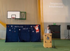 Na tle granatowego płótna z napisem Wesołych Świąt i wyżej wyświetlonego napisu Świąteczne kolędowanie uczestników zapowiada przewodniczący samorządu uczniowskiego.
