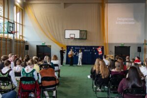 Na pierwszym planie rzędy uczniów obserwujących występ dwóch śpiewajacych uczennic i wspomagającego ich skrzypka.