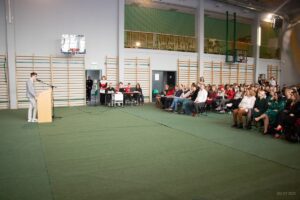 Po lewej stronie przemawiający do zebranych przewodniczący samorządu szkolnego po prawej w pierwszych rzędach nauczyciele a w dalszych uczniowie.