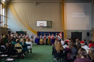 W głębi sceny przy dekoracji z napisem Wesołych Świąt ustawione w rząd dziewczęta w strojach góralskich śpiewają kolędę.