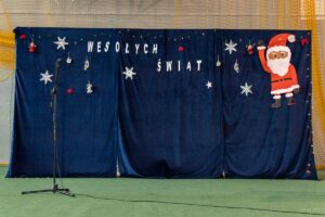 Napisy końcowe dekoracja na grantowym płótnie biały napis Wesołych Świąt i po prawej stronie radośnie machający Mikołaj.