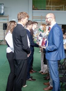 Zbliżenie na kierownictwo szkoły , żegnane przez delegację klas piątych. Na pierwszym planie pan wicedyrektor z otrzymanym prezentem pożegnalnym.