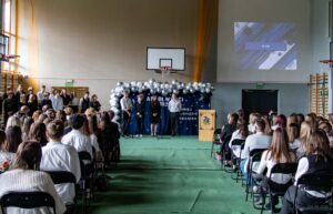 Na pierwszym planie siedzący zgromadzeni na akademii ,słuchający stojących w głębi sali i deklamujących strofy pożegnania uczniów klas IV. Po prawej stronie u góry na ścianie wyświetlają się równocześnie wspomnieniowe slajdy poszczególnych klas absolwentów.