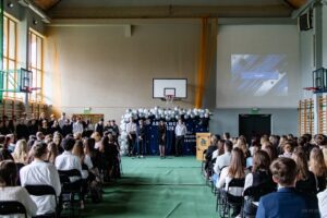 Następne zdjęcie, na którym na pierwszym planie siedzący zgromadzeni na akademii ,słuchający stojących w głębi sali i deklamujących strofy pożegnania uczniów klas IV. Po prawej stronie u góry na ścianie wyświetlają się równocześnie wspomnieniowe slajdy poszczególnych klas absolwentów.