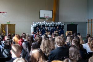 Na pierwszym planie uczniowie oglądają występ żegnającego maturzystów tria muzycznego złożonego z nauczyciela i uczennicy z gitarami i śpiewającej solistki.