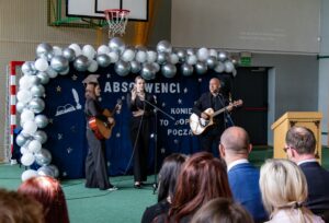 Na tle napisu Absolwenci 2026 występuje żegnające absolwentów trio muzyczne dwie uczennice i nauczyciel grający na gitarze.