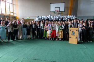 Część uczniów klas piątych stoi i radośnie macha oraz śpiewa ciesząc się ze świadectw ukończenia szkoły.