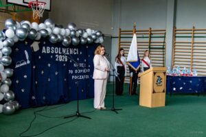 Na scenie przemawia do zebranych Pani Dyrektor za nią dekoracja i stojący poczet sztandarowy.