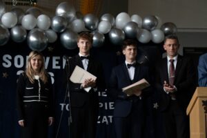 Zbliżenie na absolwentów , laureatów i zwycięzców olimpiad przedmiotowych w towarzystwie wychowawców, którzy stoją po obu bokach.