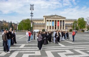 Na środku placu grupa uczniów słuchająca przewodnika, w tle budynek muzeum.