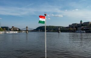 Widok na węgierską flagę w kolorach czerwony, biały i zielony zatkniętą jako bandera na statku wycieczkowym w tle jeden z budapesztańskich mostów.