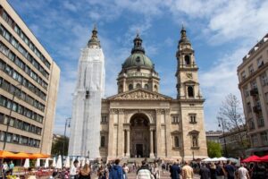 Miejski plac z turystami w tle budapesztańska kontrkatedra z dwoma wysokimi wieżami po bokach.