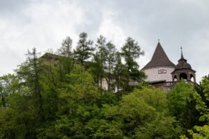 Pop prawej stronie wieża zamkowa wyłaniająca się z pośród drzew za którymi ukrywają się dalsze zabudowania zamkowe.