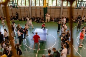Liczne grono uczniów obserwuje zacięty pojedynek tenisa stołowego między wicedyrektorem i nauczycielką.